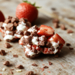 A colorful bowl of chocolate strawberry yogurt clusters with visible strawberries, coated in smooth chocolate, arranged on a white plate, surrounded by scattered strawberries and chunks of chocolate, styled casually on a wooden surface with soft natural light.